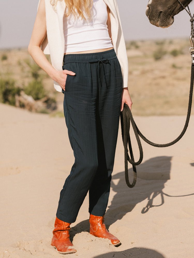 A person stands on sandy ground holding horse reins, wearing a white tank top, Cozy Earth Women's Sunset Cropped Pant, a cream sweater draped over their shoulders, and orange cowboy boots. The horse's head is partially visible on the right.|Color:Eclipse