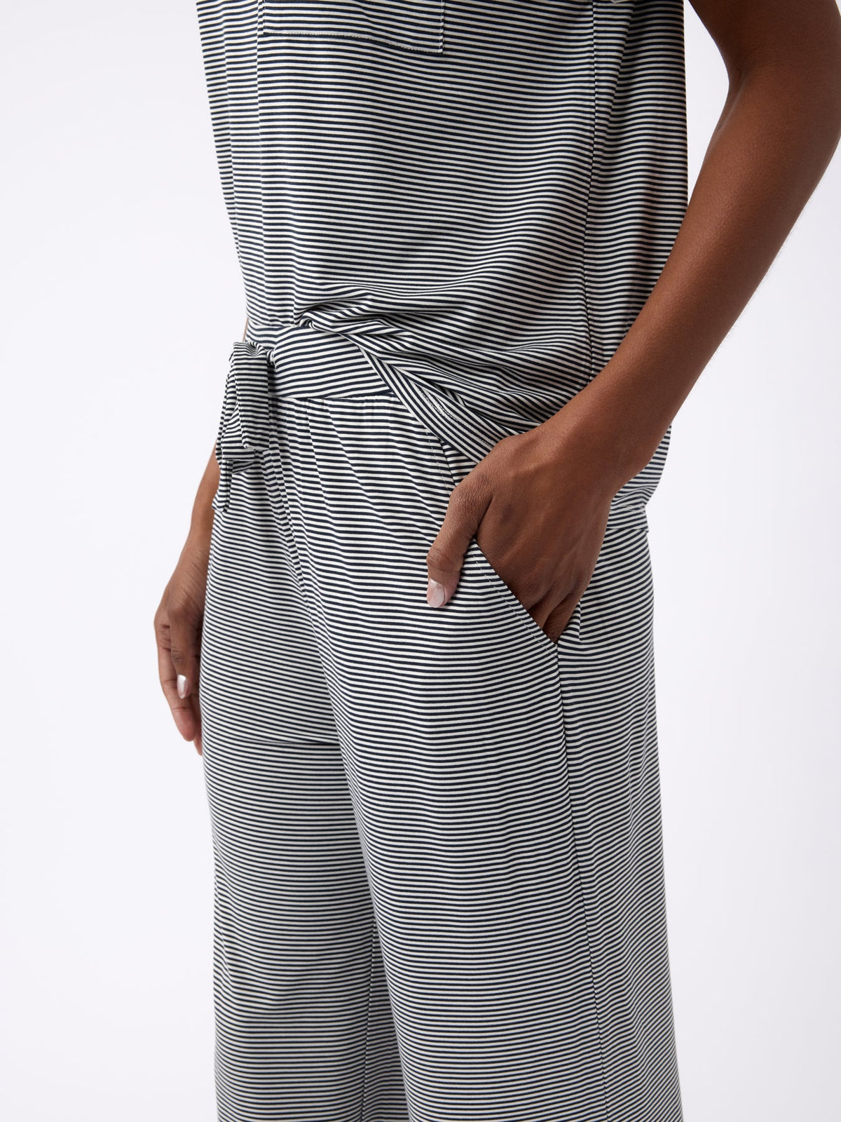 A person is wearing a Cozy Earth Women's Bamboo Stretch-Knit Pant, featuring a gray striped design, short-sleeved pocket shirt, and pants with a waist tie detail. Their hand rests in the pant pocket. |Color:Eclipse Mini Stripe