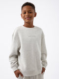 A smiling boy wearing the Kid's CityScape Crewneck by Cozy Earth, featuring "COZY EARTH" printed on it, stands against a plain white background. |Color:Heather Grey