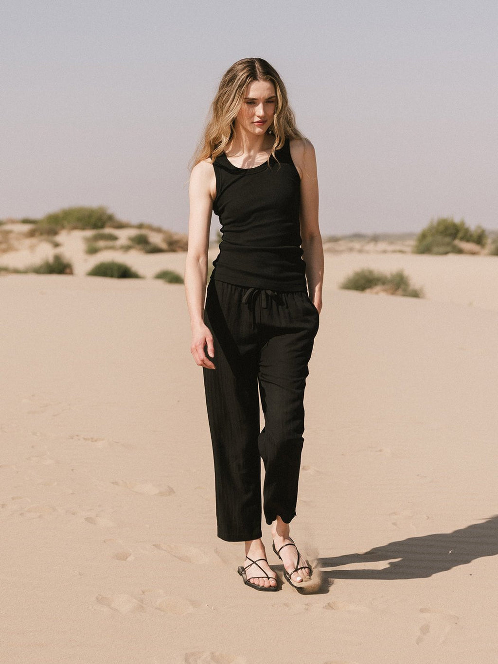 A woman with long blonde hair, wearing Cozy Earth's Women's Sunset Cropped Pant and a black sleeveless top, strolls on sandy terrain with sparse shrubs under a clear sky. |Color:Jet Black