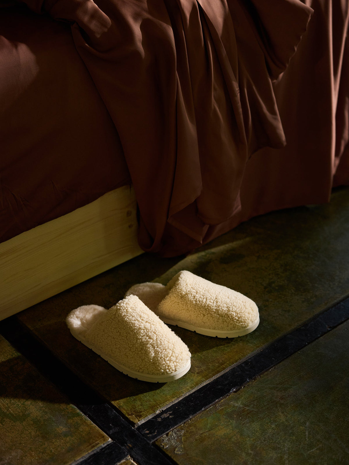 A pair of Cozy Earth's Puffy Sheep Slippers, in a cozy, cream-colored, fluffy fabric, lie on a mottled green floor, partially shaded. They are positioned next to a tan wooden bed frame with dark brown bedding draped over the side. The scene appears warm and inviting, suggestive of a cozy home environment. |Color:Natural