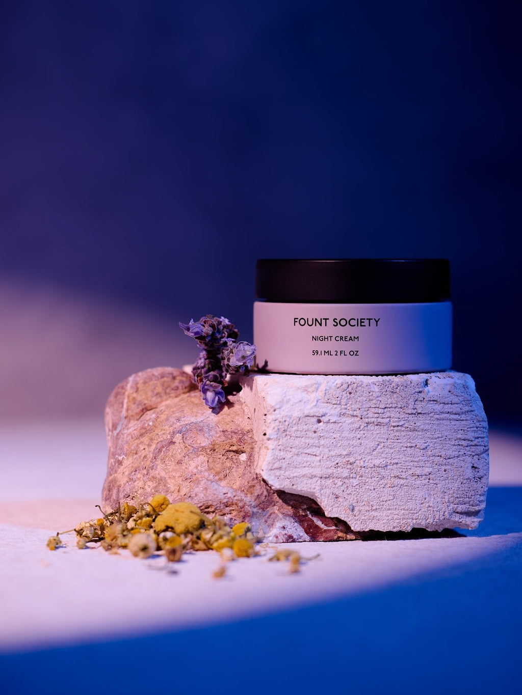 A jar of Cozy Earth Night Cream rests on a textured stone, surrounded by small dried flowers under gentle purple and pink lighting. |Size:Full Size
