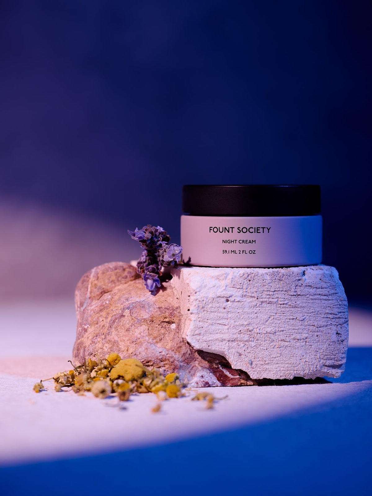 A jar of Cozy Earth Night Cream rests on a textured stone, surrounded by small dried flowers under gentle purple and pink lighting. |Size:Full Size
