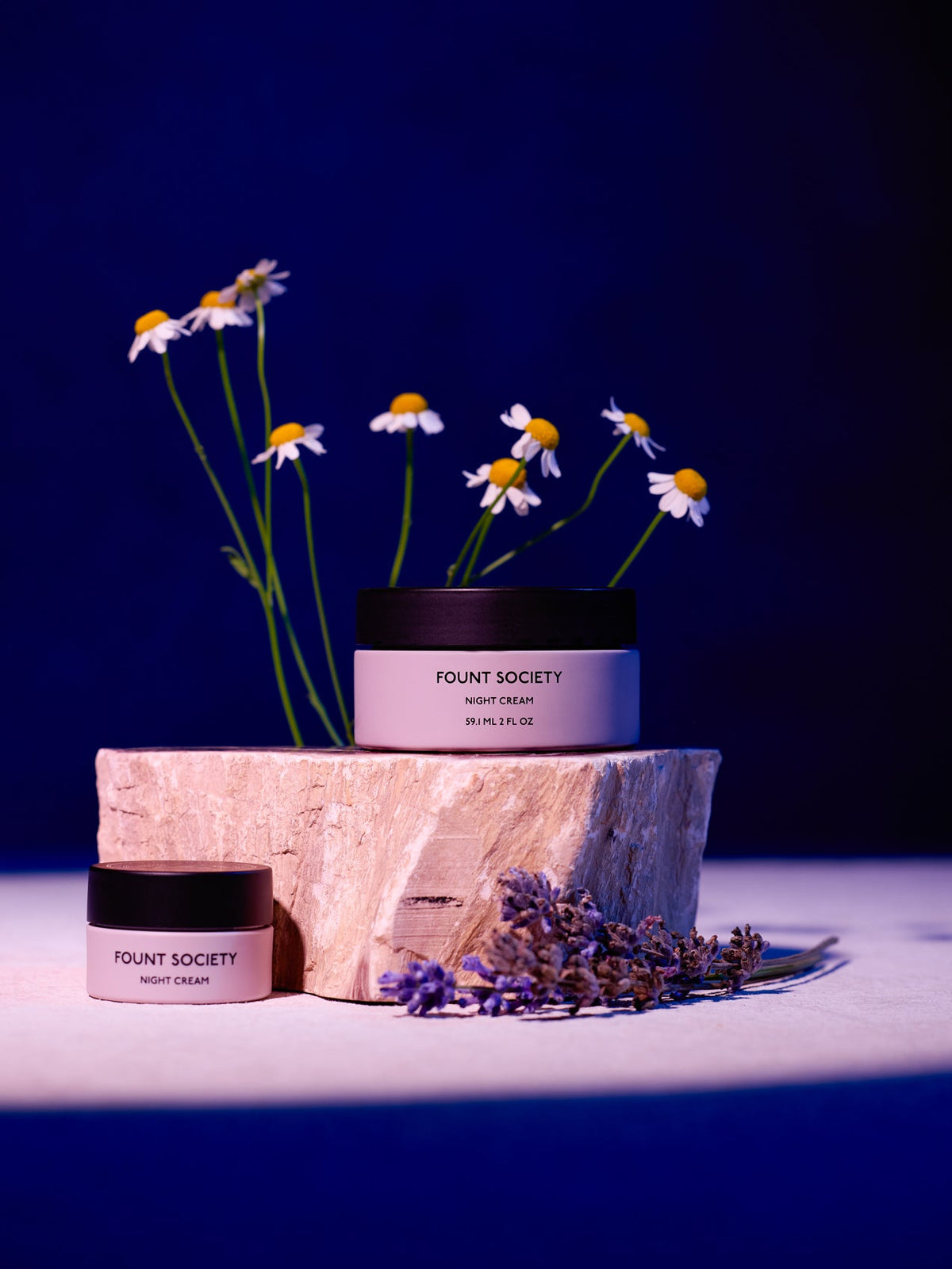 Two jars of Cozy Earth Night Cream sit on a wooden stump with a deep blue backdrop, surrounded by white daisies and purple flowers that enhance the product's natural elegance. |Size:Mini
