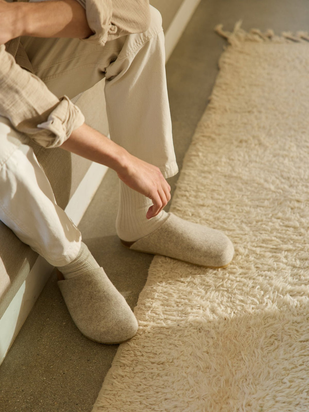 A person dressed in cream-colored loungewear and Cozy Earth's Lakehouse Clog sits on a couch's edge. Their hand rests on their thigh, and a textured cream rug covers the floor while natural light casts soft shadows across the scene. |Color:Oat
