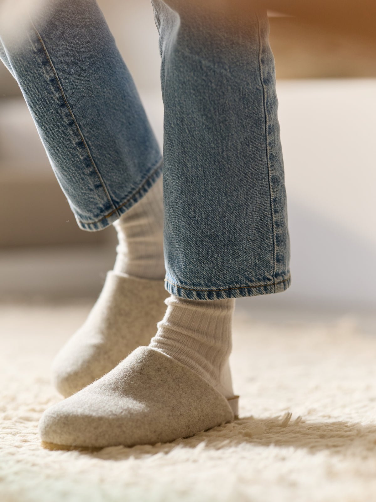 A person in blue jeans and white socks stands on a cream-colored textured rug, wearing Lakehouse Clogs by Cozy Earth. |Color:Oat