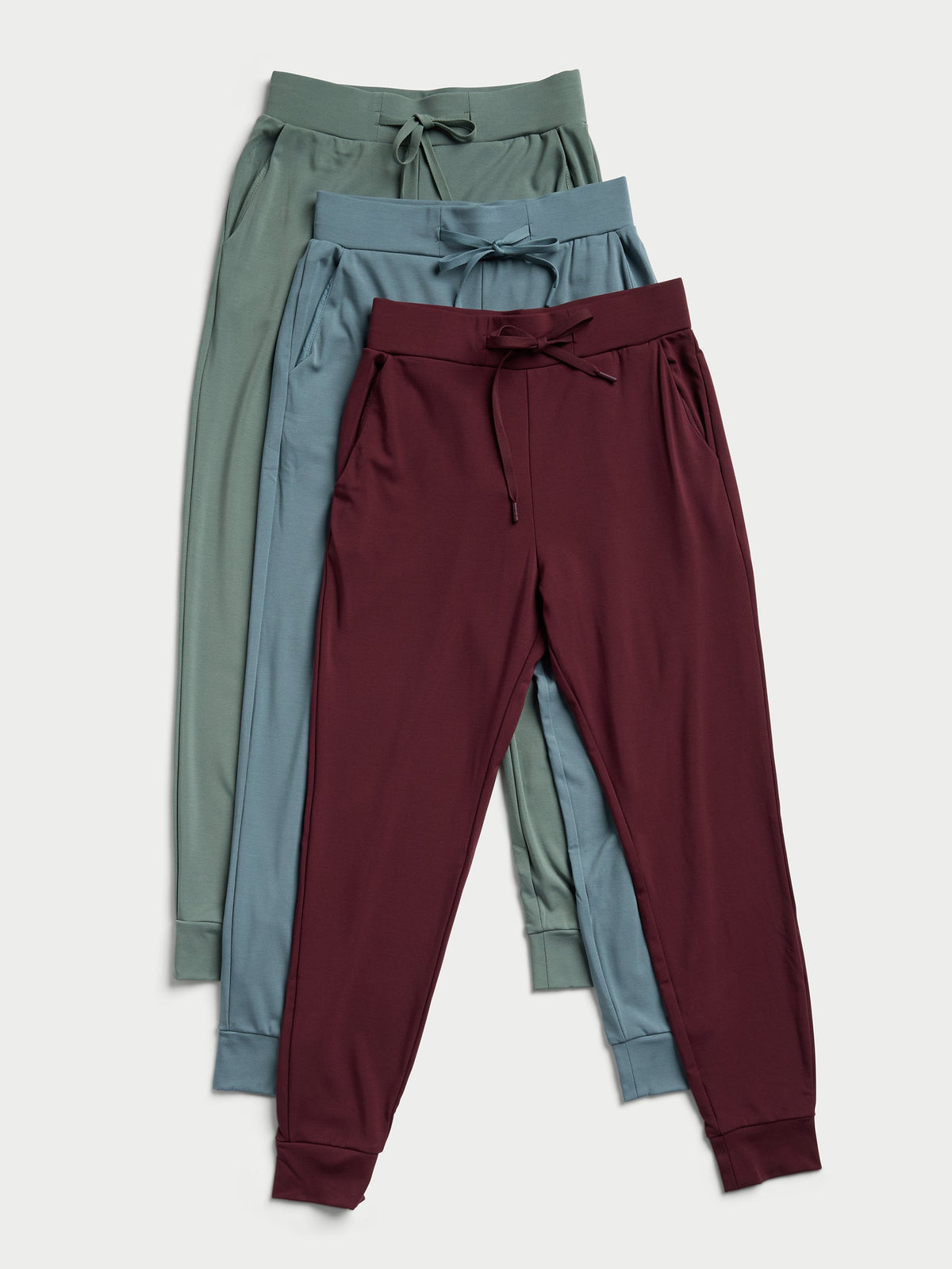 Three pairs of the Women's Bamboo Jogger Pant by Cozy Earth, in green, blue, and maroon, are placed overlapping each other. Each pair showcases a drawstring waist and cuffed ankles against a light background. |Color:Oceanside