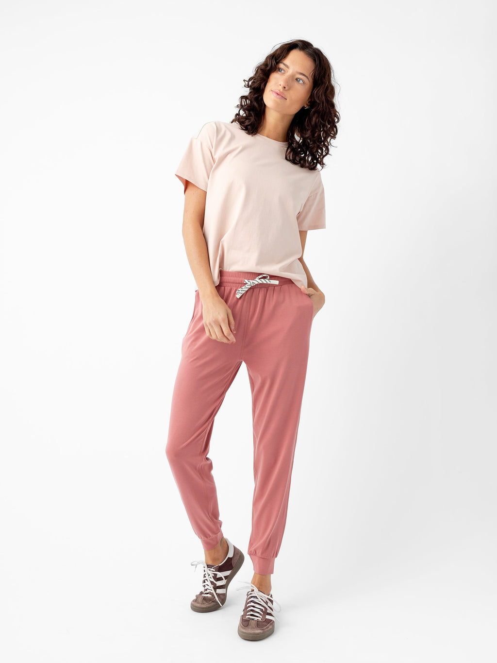 A person with curly hair is standing against a plain white background, wearing a light pink t-shirt and Women's Studio Jogger pants by Cozy Earth, tied at the waist with a white drawstring. They are also wearing brown sneakers and have their hands in their pockets, gazing slightly upwards. |Color:Rouge