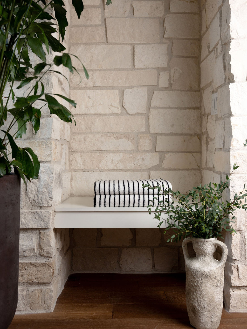 A cozy nook with a built-in white bench against a textured stone wall, featuring Luxe Bath Towels by Cozy Earth in a black and white stripe pattern. The setting is accentuated by lush green plants in pots on the wooden floor, creating a serene and inviting atmosphere. |Color:Black Stripe