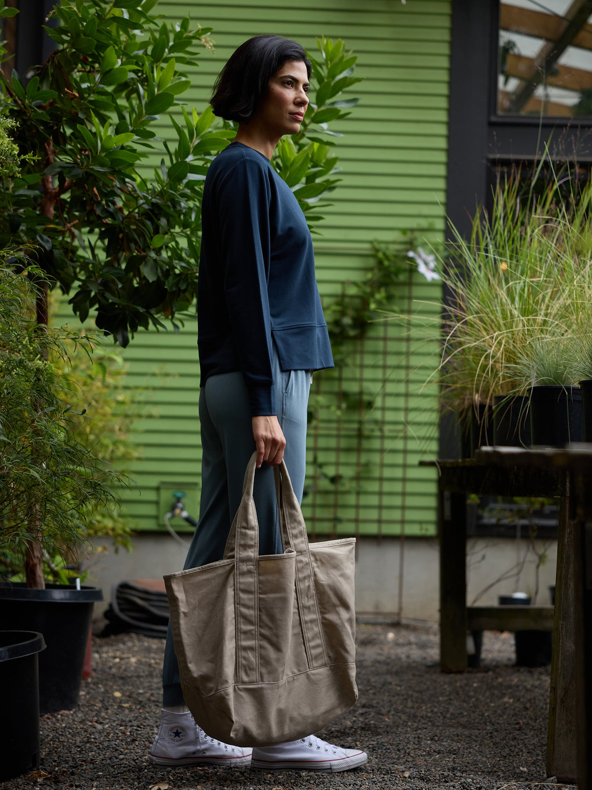 A person in a navy sweater and light blue pants stands in a garden area holding a large Waxed Canvas Tote by Cozy Earth. They have short dark hair and are wearing white sneakers. The background features green plants, trees, and a green building with horizontal siding. |Color:Taupe