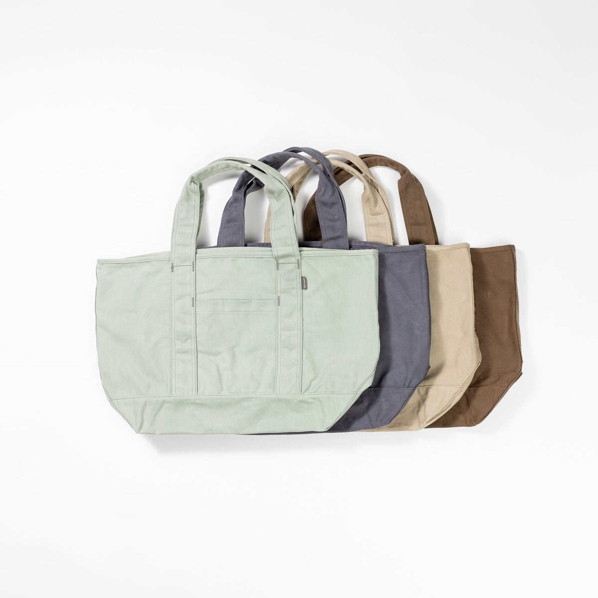 Five Waxed Canvas Tote bags from Cozy Earth are displayed in a row against a white background. The bags are arranged in descending order from left to right by color: light green, dark gray, light gray, beige, and brown. Each bag features two handles and a front pocket. |Color:Taupe
