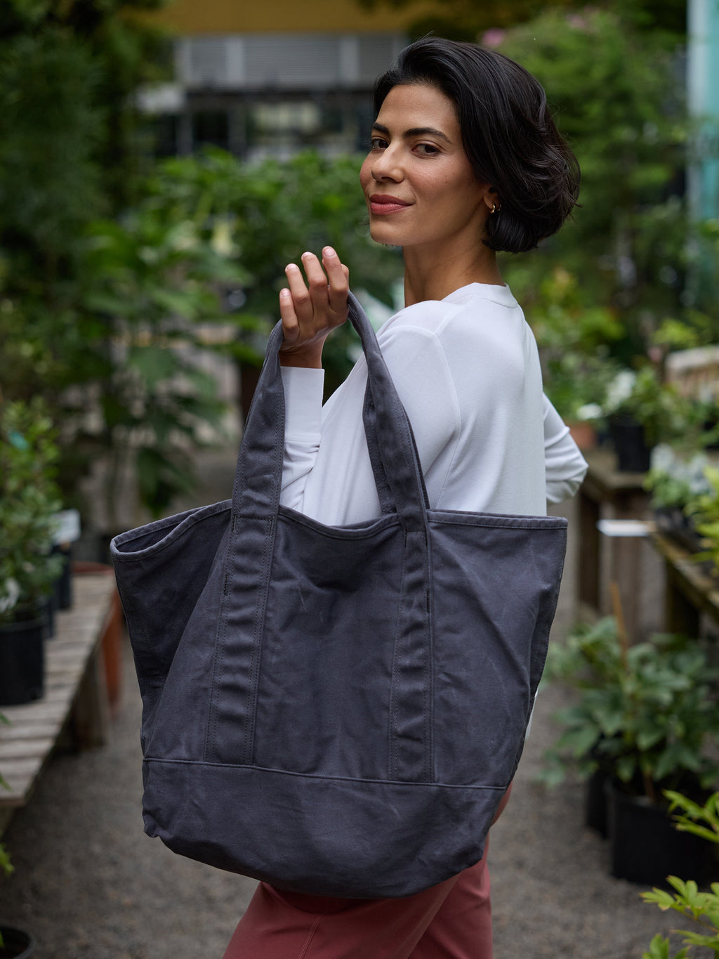 A woman with short dark hair, wearing a white long-sleeve shirt and pink pants, is standing in a lush garden center. She is smiling and looking over her shoulder while holding a large Waxed Canvas Tote from Cozy Earth. Various potted plants and greenery surround her. |Color:Washed Navy