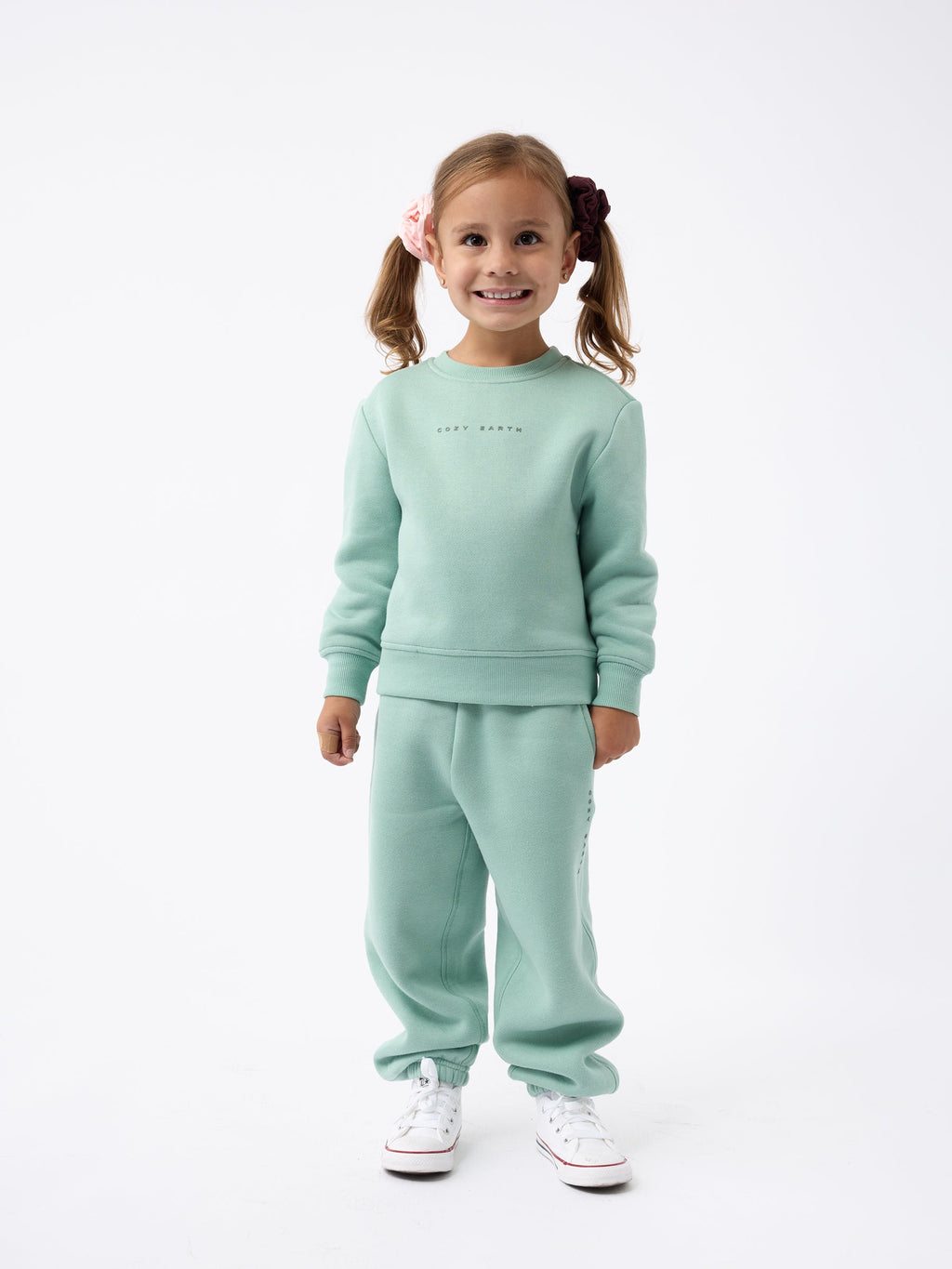 A young girl with pigtails stands smiling, dressed in the Kid's CityScape Crewneck by Cozy Earth and matching pants, paired with white sneakers against a plain white background. |Color:Wintergreen