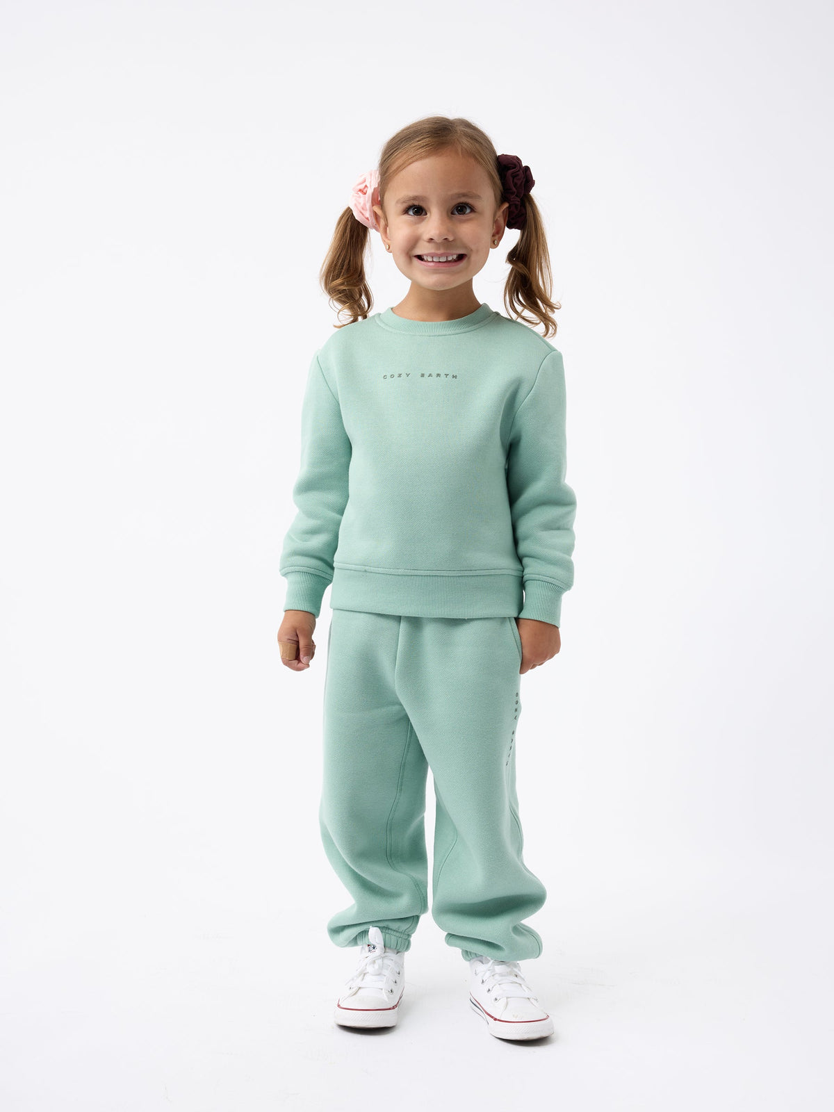 A young girl with pigtails stands smiling, dressed in the Kid's CityScape Crewneck by Cozy Earth and matching pants, paired with white sneakers against a plain white background. |Color:Wintergreen