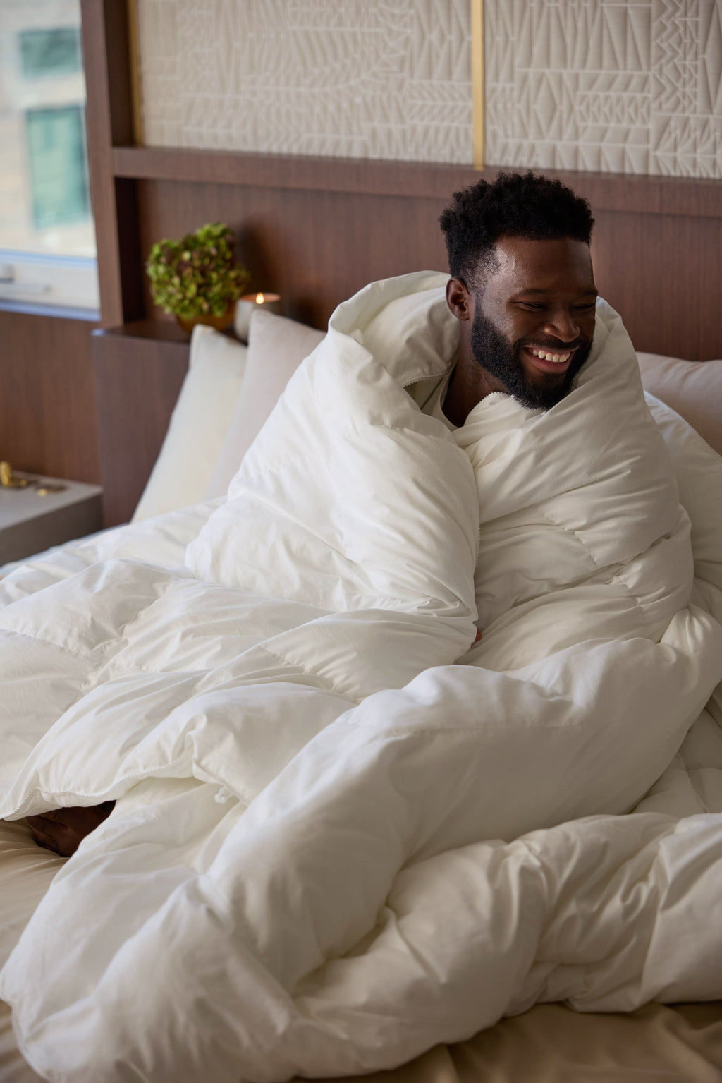 A man sits on a bed wrapped in the fluffy Cozy Earth Down Alternative Comforter, smiling and looking to the side in a cozy, well-lit bedroom. |Filling:Standard