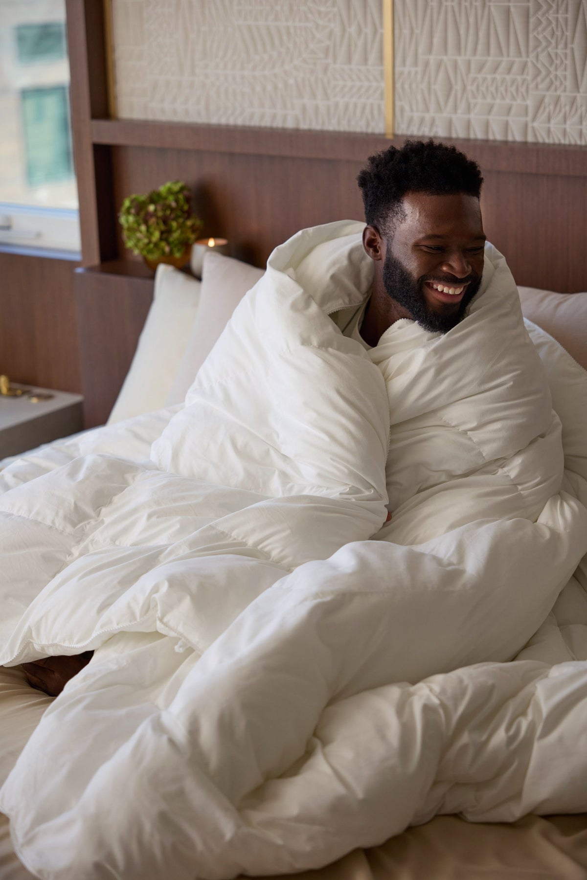 A man sits on a bed wrapped in the fluffy Cozy Earth Down Alternative Comforter, smiling and looking to the side in a cozy, well-lit bedroom. |Filling:Standard