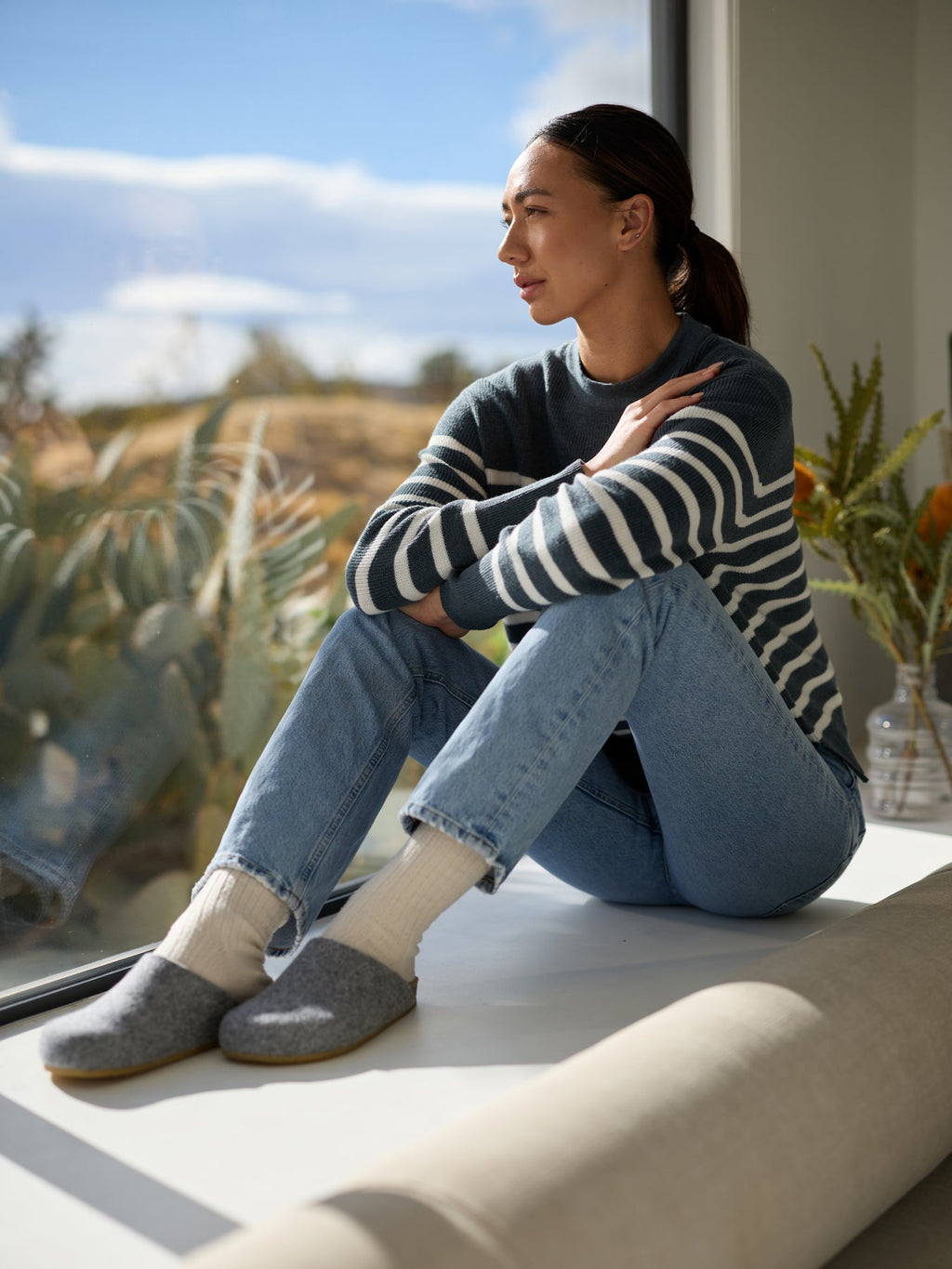 A person sits on a window seat, gazing outside thoughtfully. They are wearing a striped sweater, jeans, and Cozy Earth's Lakehouse Clog slippers. Sunlight illuminates the scene, with plants and sky in the background. |Color:Charcoal