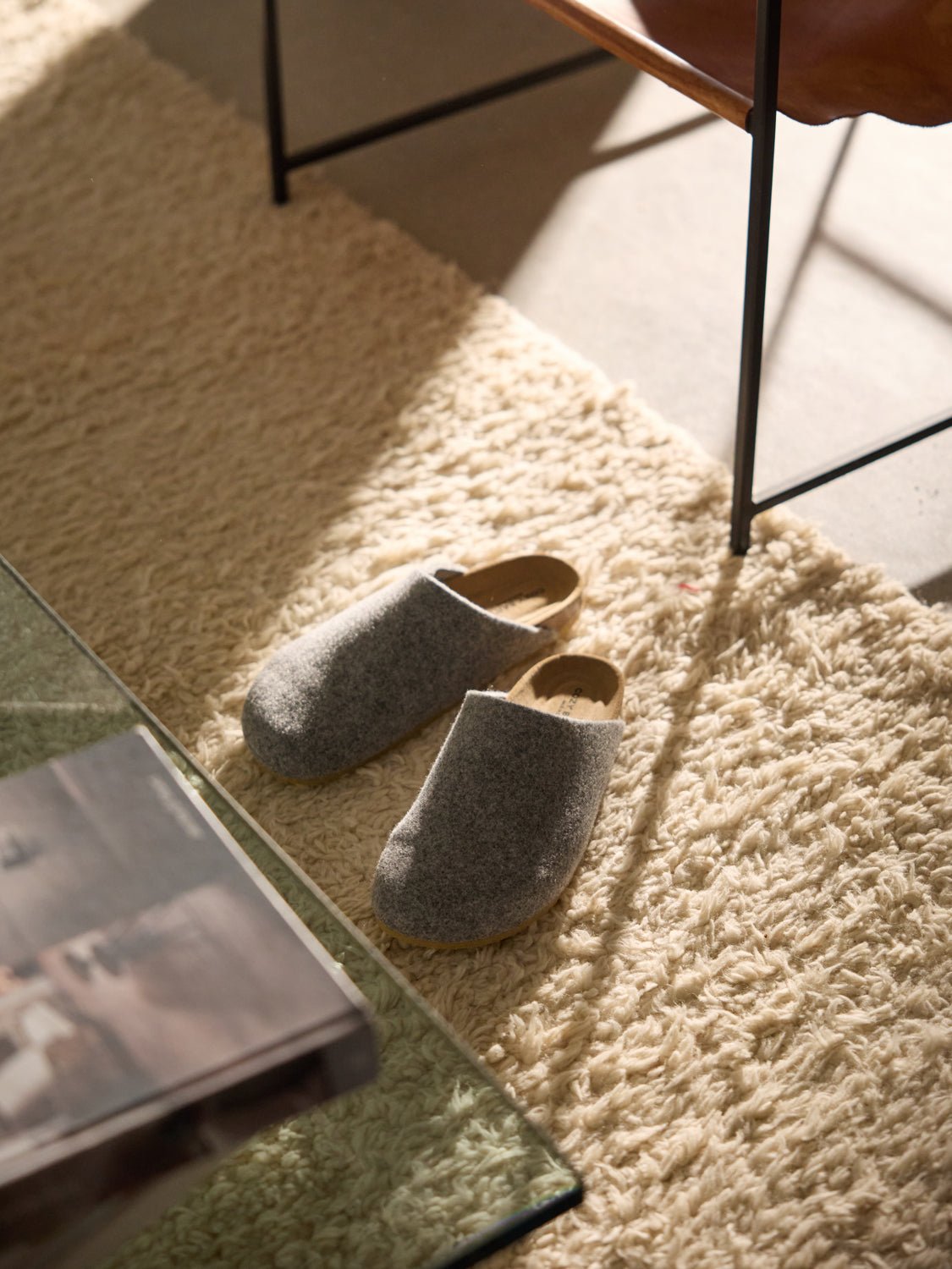 A pair of Cozy Earth's Lakehouse Clog wool slippers rests on a soft, cream-colored shag rug near a glass coffee table and leather chair. Sunlight from a nearby window casts warm shadows across the scene. |Color:Charcoal