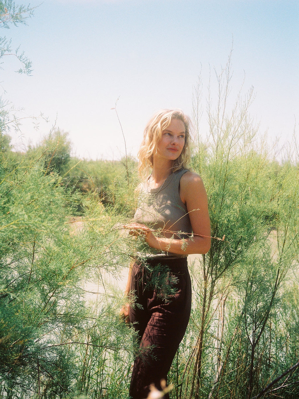 A person with shoulder-length blonde hair stands amidst tall, green, wispy plants. They are wearing a gray sleeveless top and the Women's Sunset Cropped Pant by Cozy Earth, gazing to the side. A bright, clear sky and lush greenery are visible in the background. |Color:Cacao