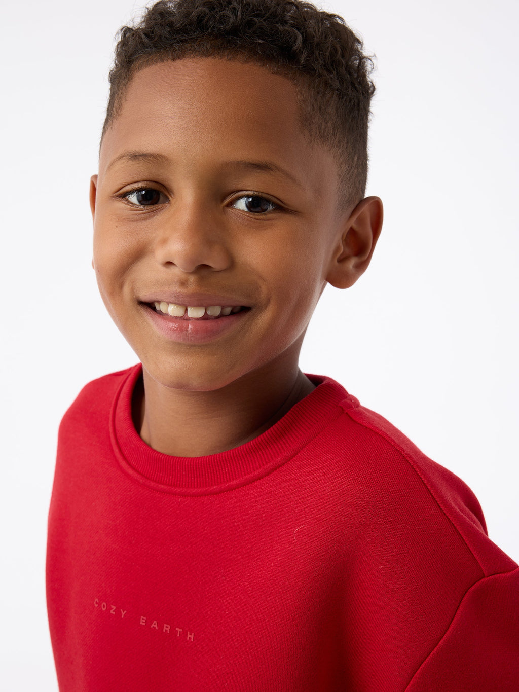 A young boy, dressed in a Cozy Earth Kid's CityScape Crewneck, smiles warmly at the camera against a white background. |Color:Crimson