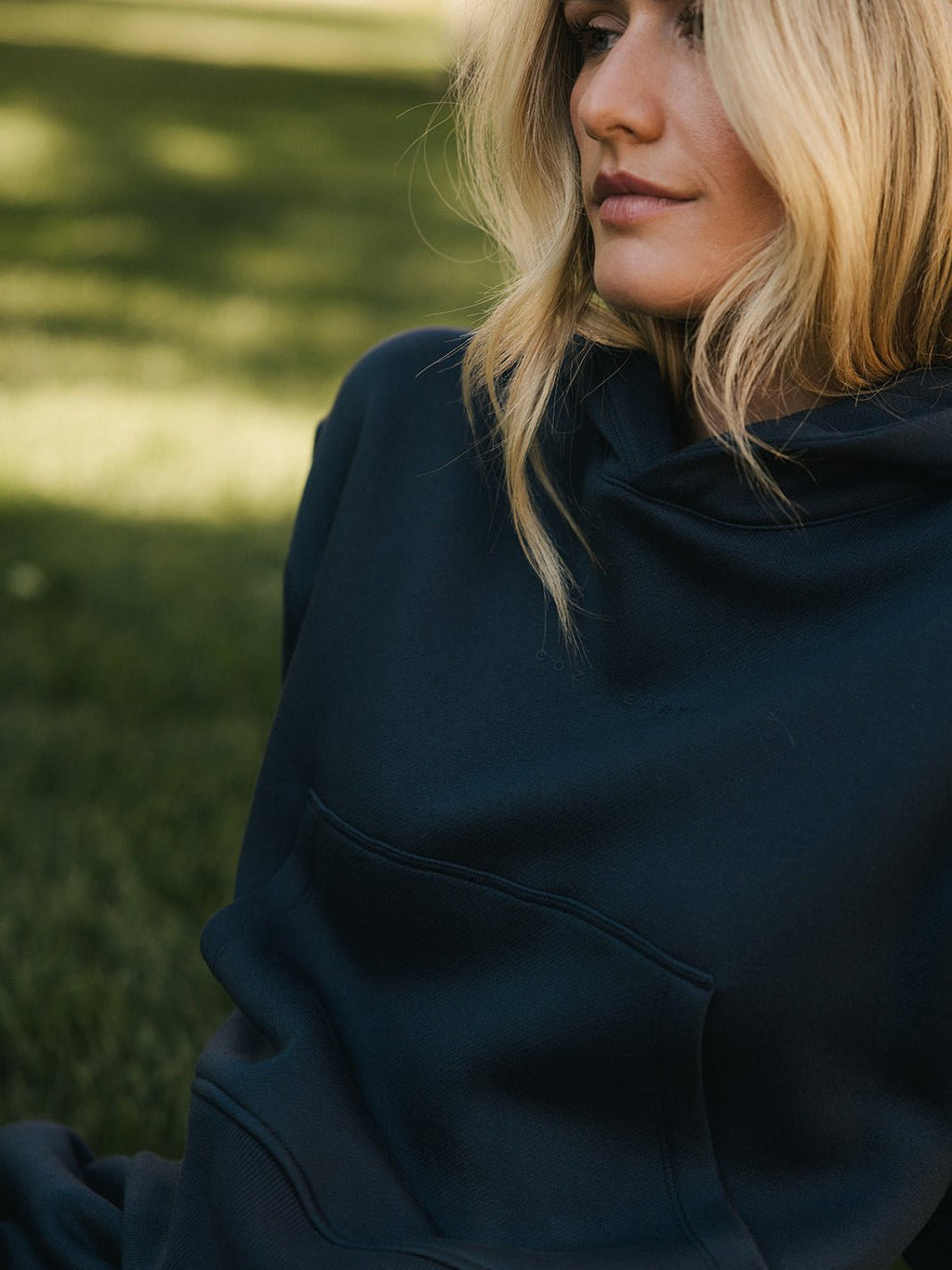 A person with long blonde hair in a Women's CityScape Cropped Hoodie by Cozy Earth is sitting outdoors on the grass. Their face is partially visible, and they appear to be relaxed, enjoying the serene environment. The background is blurred, highlighting a green, grassy area. |Color:Eclipse