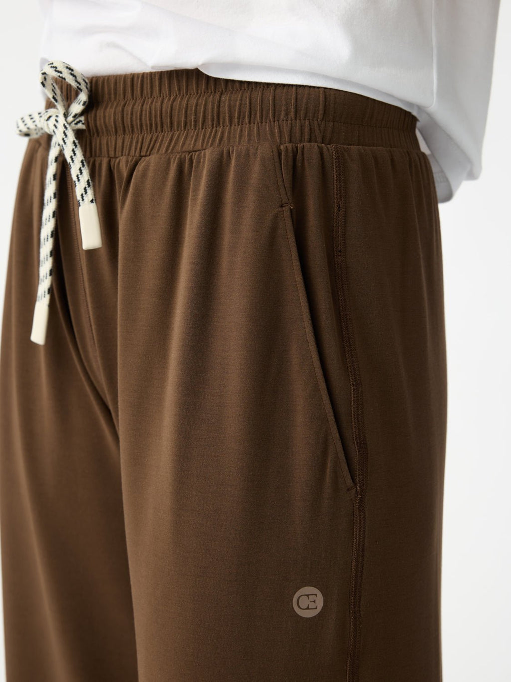 A person wearing Cozy Earth's Women's Studio Cropped Wide Leg Pant in brown, featuring a white-and-black drawstring and a small emblem near the pocket, paired with a white top. |Color:Espresso