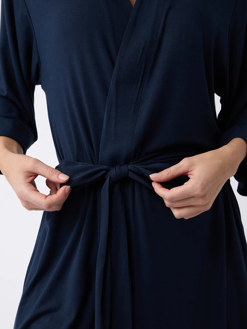 An individual secures the belt of a Cozy Earth Women's Stretch-Knit Bamboo Kimono Robe in dark blue, highlighting their hands and the soft, long-sleeved fabric. |Color:Navy