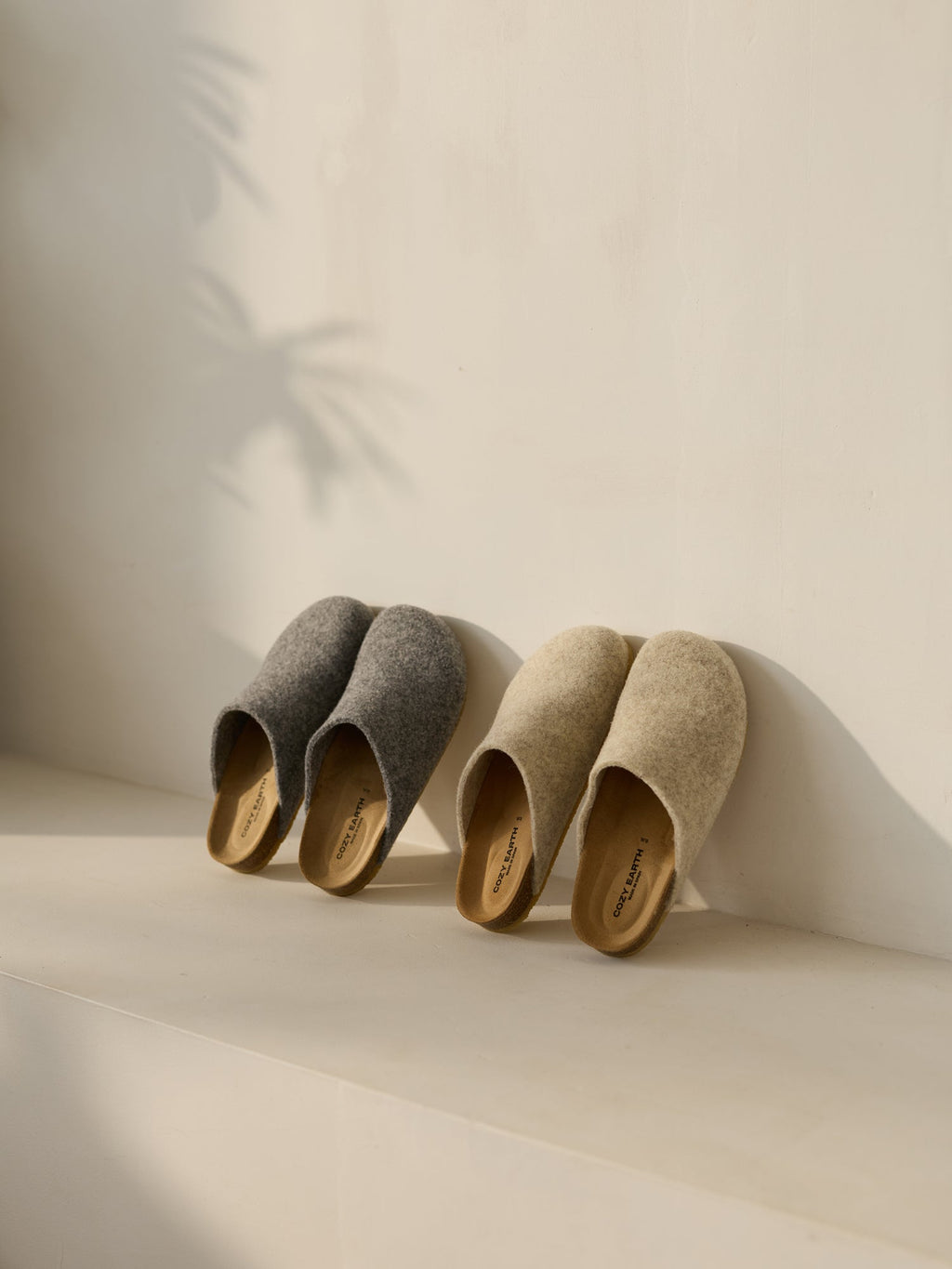 Two pairs of Cozy Earth's Lakehouse Clogs sit on a light surface. One is gray, the other beige, with soft sunlight casting gentle shadows of leaves on the wall behind them, creating a calm and cozy ambiance.|Color:Oat