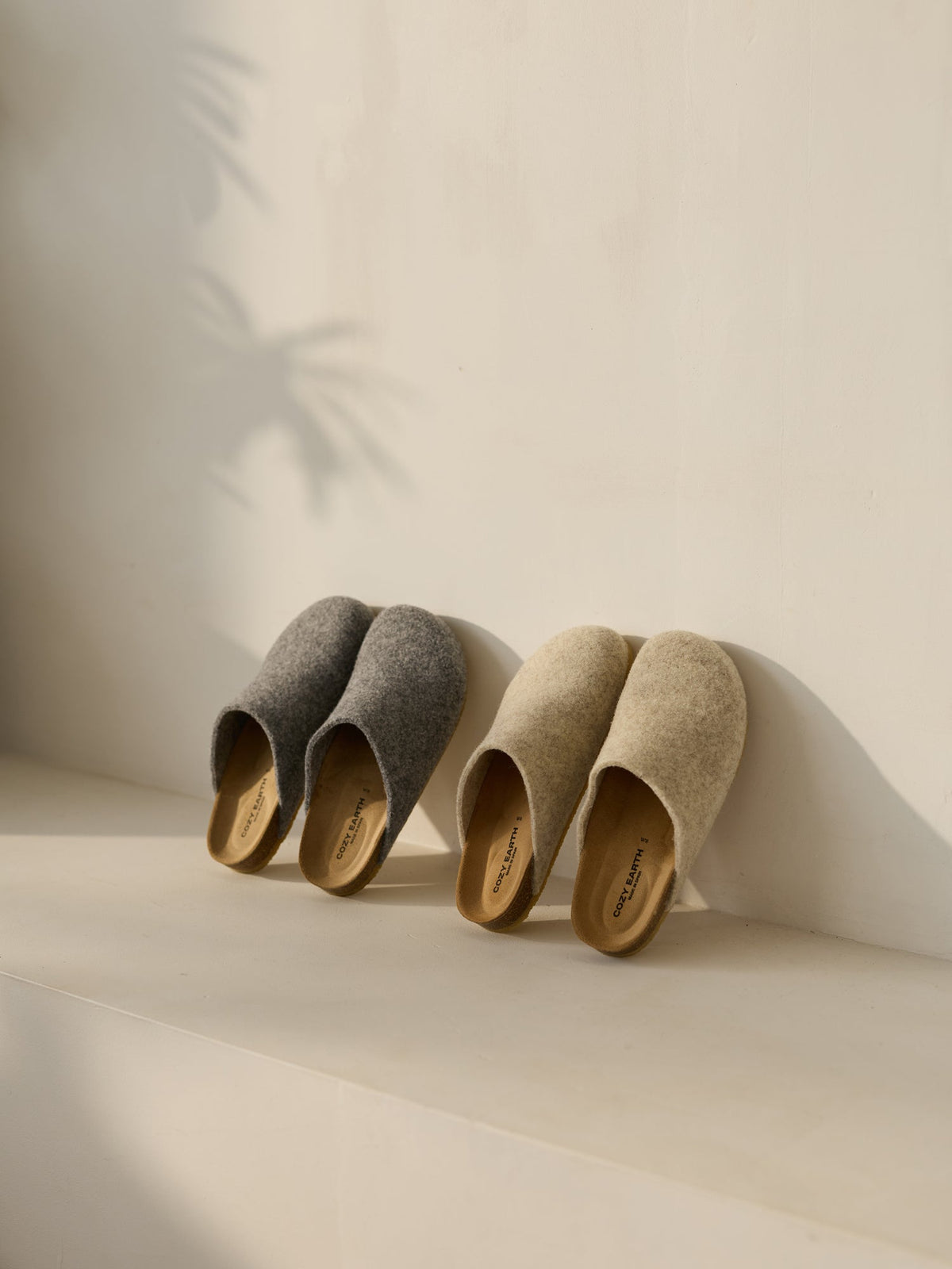 Two pairs of Cozy Earth's Lakehouse Clogs sit on a light surface. One is gray, the other beige, with soft sunlight casting gentle shadows of leaves on the wall behind them, creating a calm and cozy ambiance.|Color:Oat