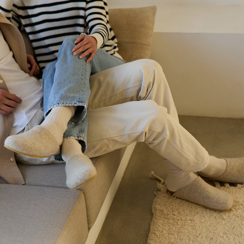 Two people relax on a beige sofa in casual, light-colored clothing. One in striped tops and jeans sits across the other's lap. Both wear Cozy Earth's Lakehouse Clog wool slippers, creating a cozy atmosphere. |Color:Oat