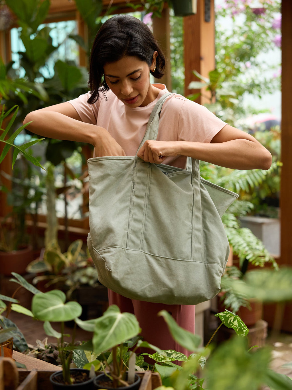 A person wearing a light pink shirt inspects the contents of a large Waxed Canvas Tote by Cozy Earth, which is light green in color. They are standing in a lush, indoor garden filled with various green plants. The ambiance is serene and filled with natural light from large windows. |Color:Palm