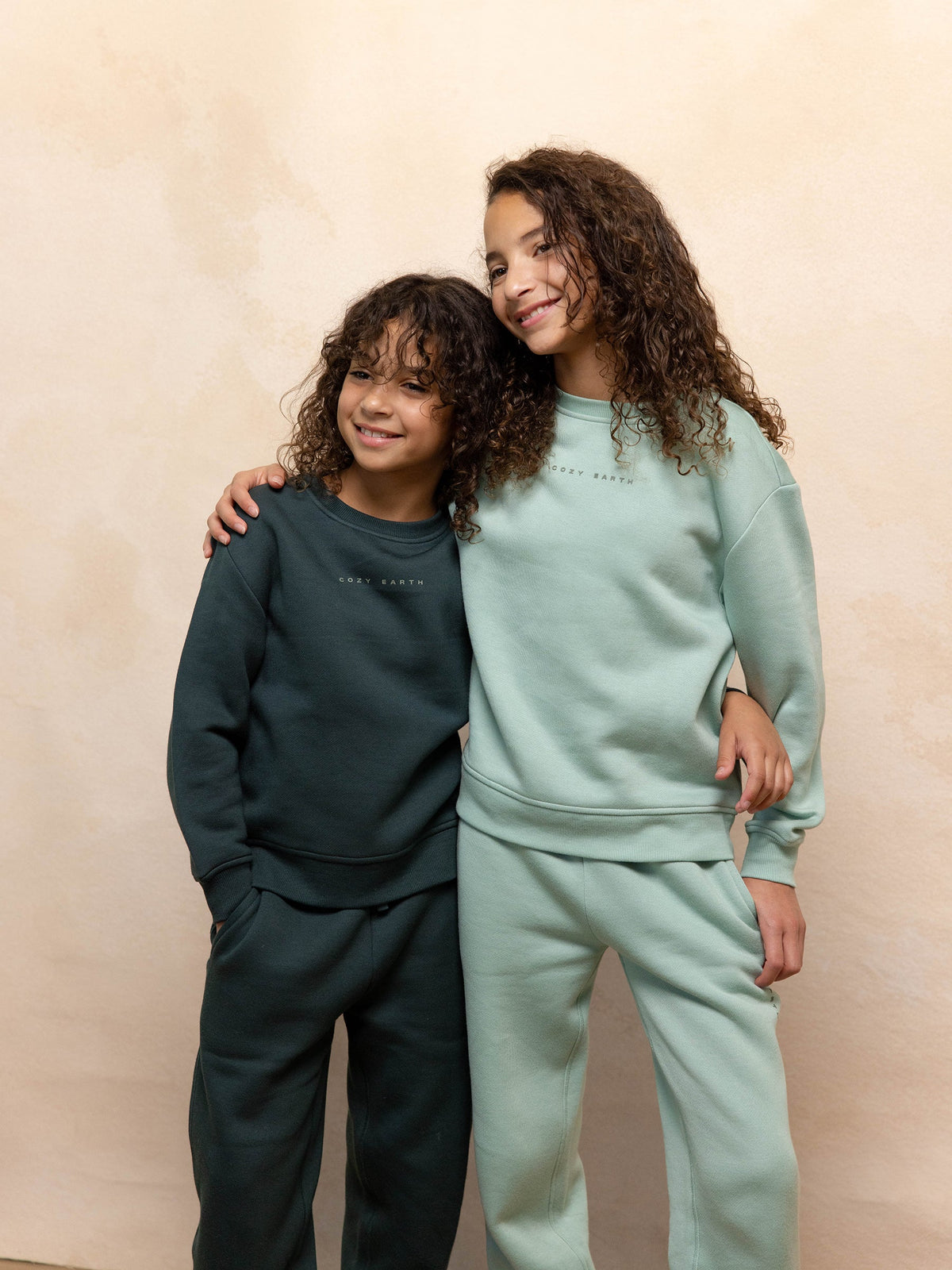 Two children with curly hair, both wearing matching outfits from Cozy Earth's Kid's CityScape Crewneck collection, smile and stand side by side with their arms around each other. One child is dressed in a dark green tracksuit, while the other wears a light green tracksuit against a neutral-toned background. |Color:Spruce