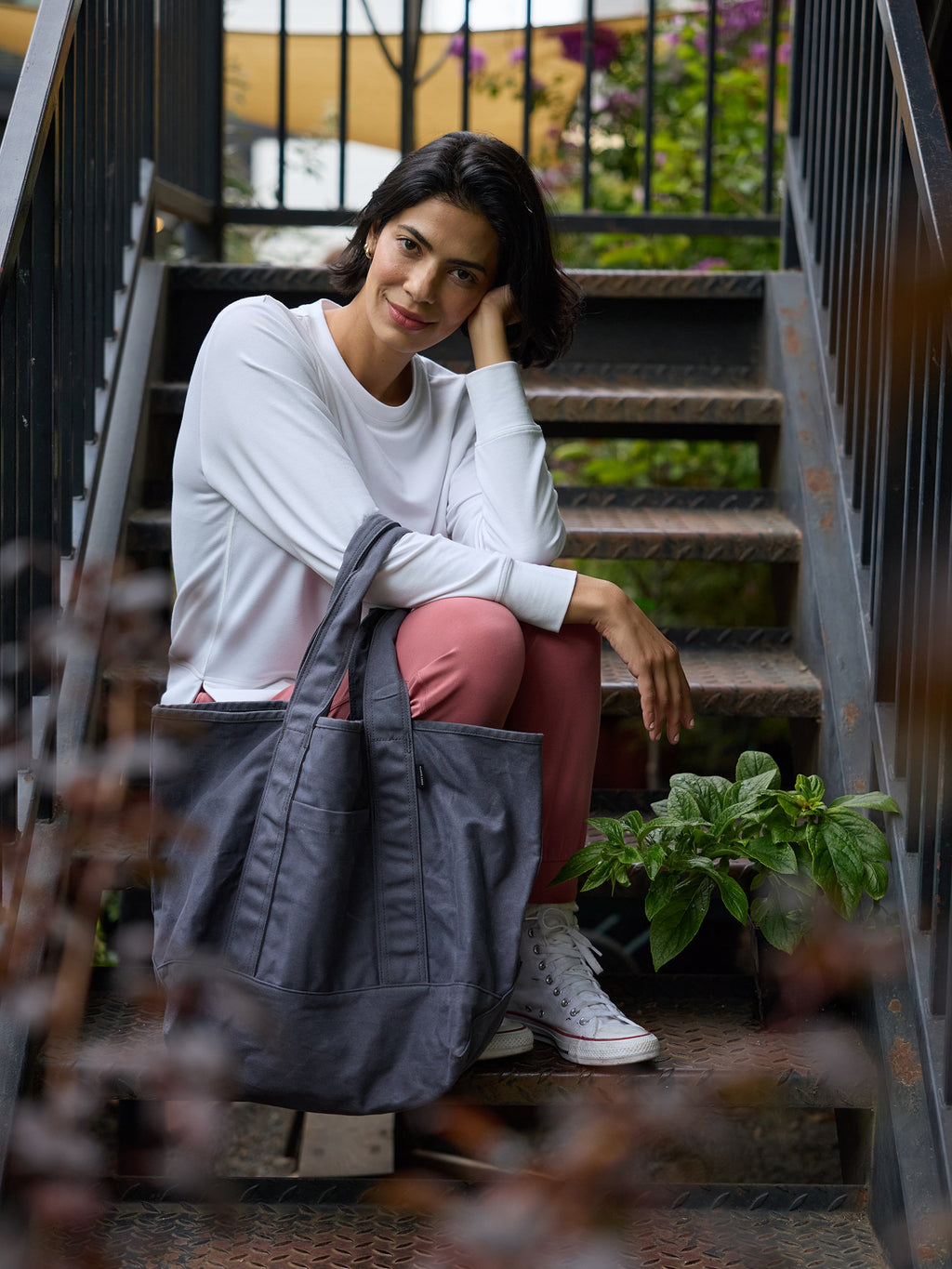 A woman with short, dark hair sits on metal stairs outdoors, resting her head on her hand. She is wearing a white sweater, pink pants, and white sneakers, and is holding a large Waxed Canvas Tote from Cozy Earth. Green foliage is visible in the background. |Color:Washed Navy