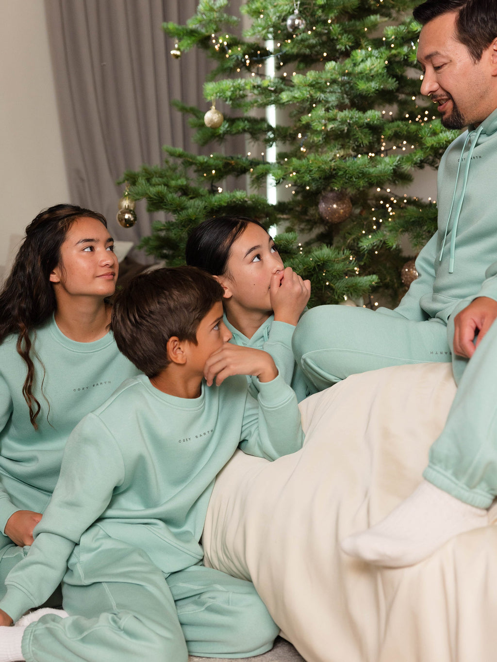 A family sitting together in matching Cozy Earth Kid's CityScape Crewnecks beside a decorated Christmas tree. The children are looking attentively at a man who is speaking, creating a cozy and festive atmosphere. |Color:Wintergreen