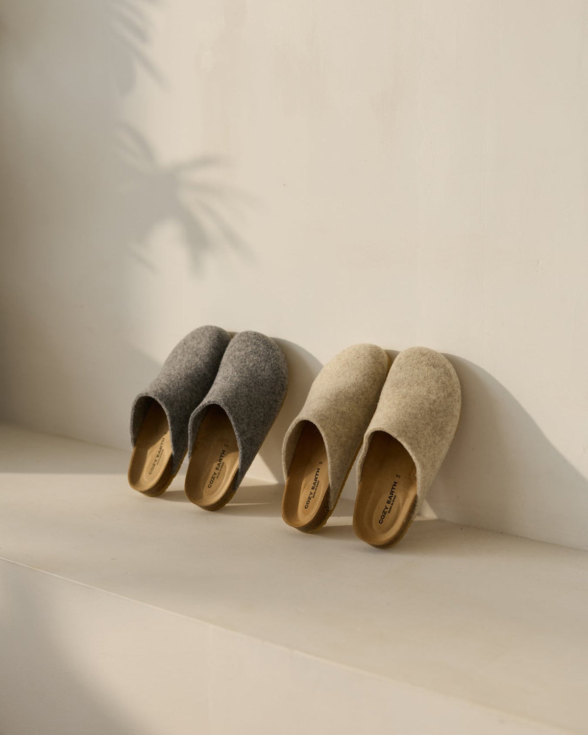 Two pairs of Cozy Earth's Lakehouse Clog slippers, one gray and one beige, with wooden soles and a minimalist design, are placed side by side on a light surface. A soft shadow of a plant appears on the wall. |Color:Charcoal