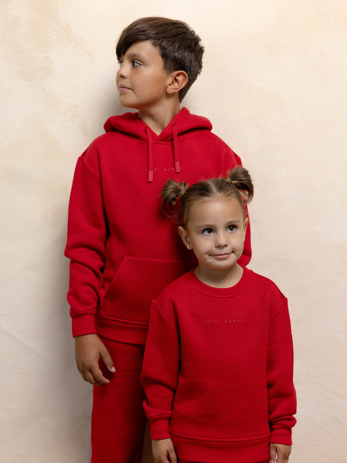 Two children in matching red Kid's CityScape Crewnecks by Cozy Earth stand in front of a beige wall. The older child, a boy, looks to the side while the younger child, a girl with pigtails, smiles at the camera. |Color:Crimson