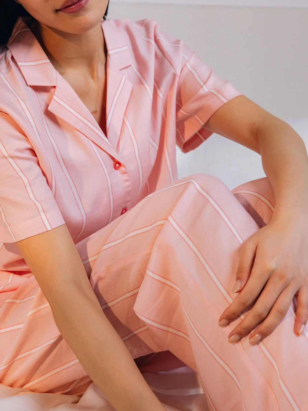A person in pink striped pajamas sits on a bed with arms resting on their knees, showcasing the Women's Soft Woven Pajama Pant from Cozy Earth, featuring a relaxed fit and button-up shirt. The background is softly blurred. |Color:Sweetheart Stripe