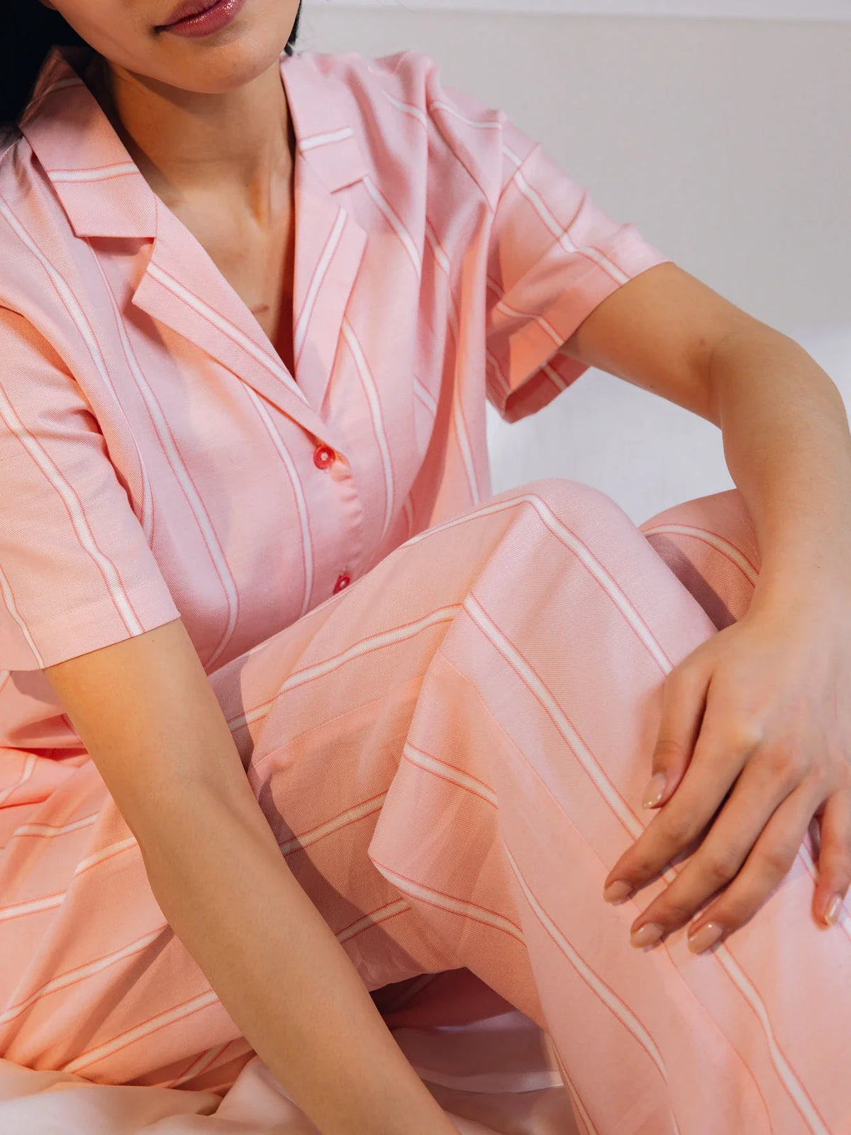 A person in pink striped pajamas sits on a bed with arms resting on their knees, showcasing the Women's Soft Woven Pajama Pant from Cozy Earth, featuring a relaxed fit and button-up shirt. The background is softly blurred. |Color:Sweetheart Stripe