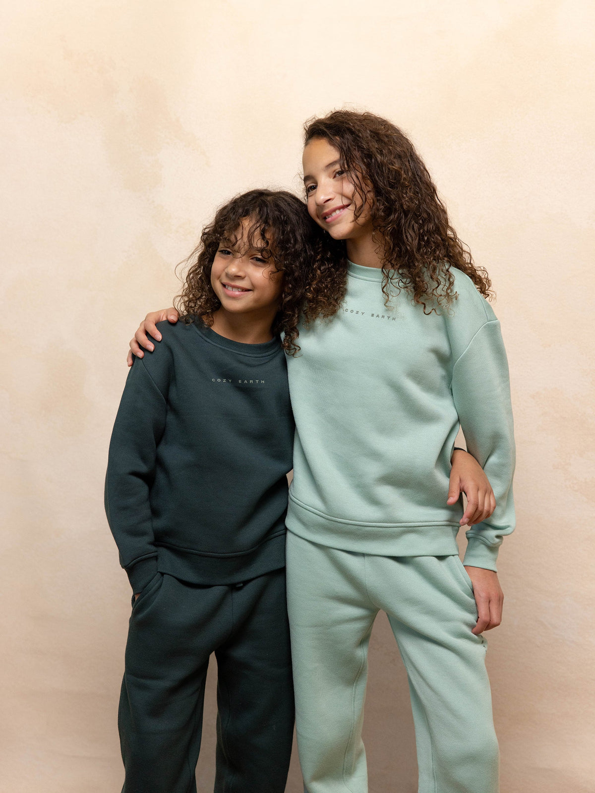 Two smiling children with curly hair are wearing Cozy Earth's Kid's CityScape Crewneck sweatshirts and pants. One is dressed in a dark green set while the other sports a light green ensemble. They pose affectionately against a neutral backdrop, with the older child wrapping an arm around the younger sibling. |Color:Wintergreen