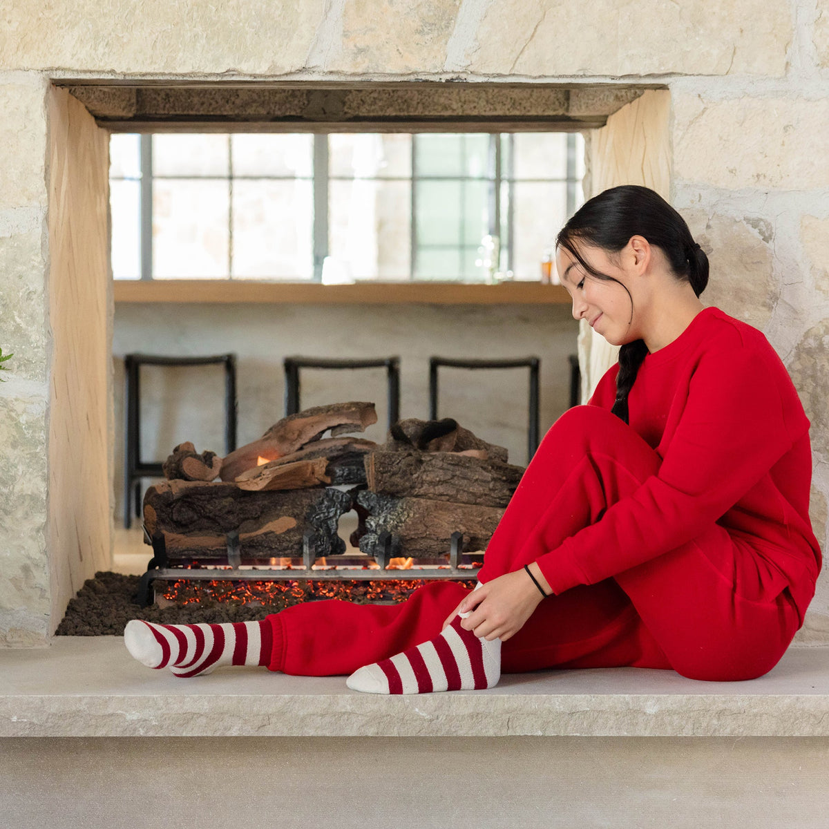 A child wearing Cozy Earth's Kid's CityScape Crewneck and striped socks sits by a stone fireplace, adjusting their sock. The setting is cozy, with logs in the fireplace and a reflective window above. |Color:Crimson