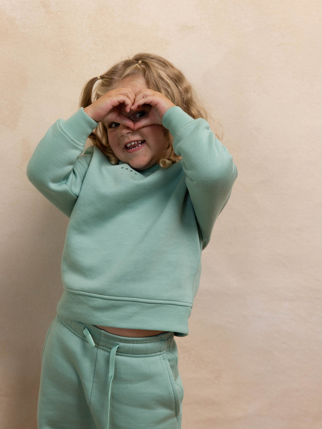A young child with curly blonde hair wearing Cozy Earth's Kid's CityScape Crewneck in mint green forms a heart shape with their hands over their face. They are smiling and standing against a beige background. |Color:Wintergreen