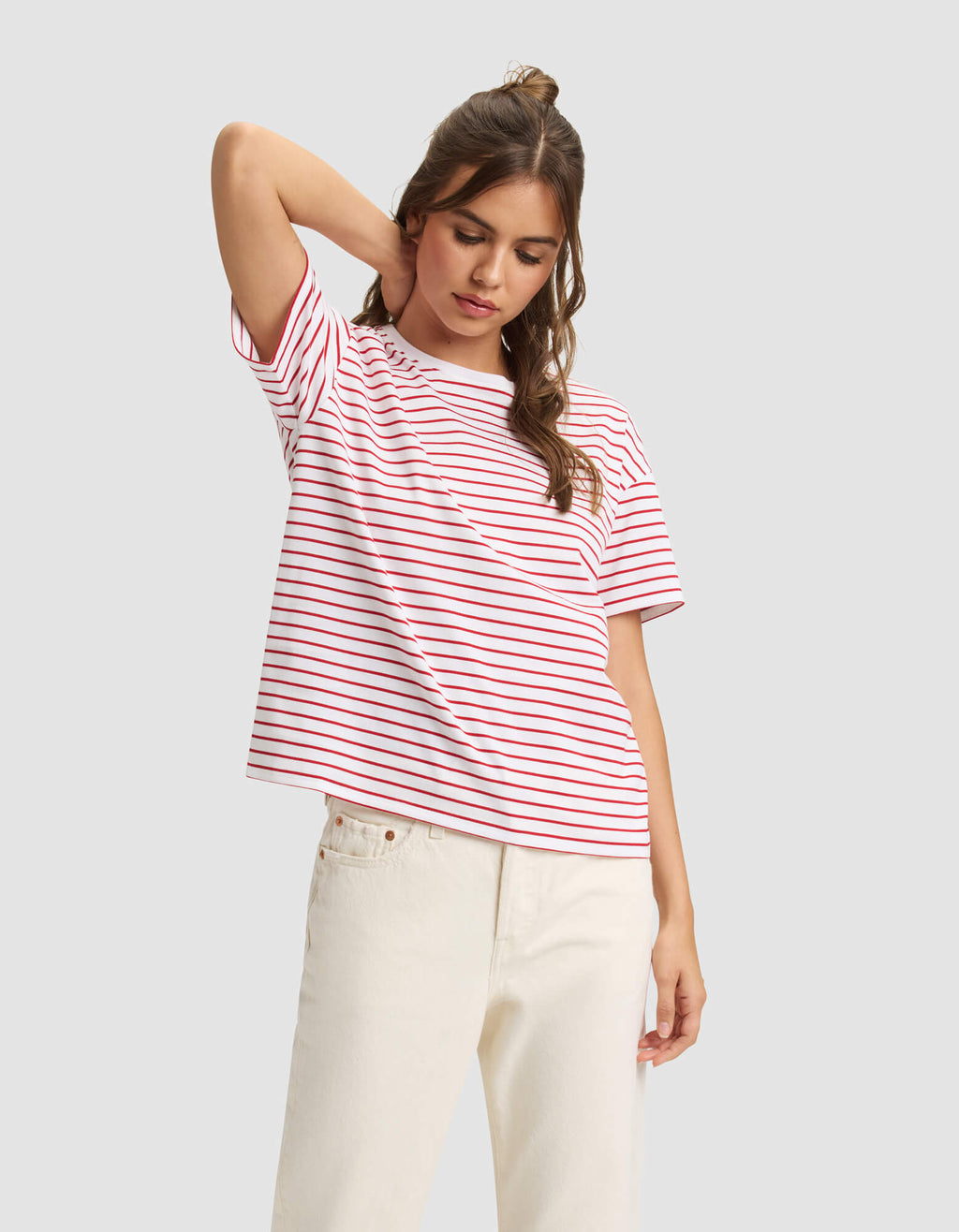 A woman models the Cozy Earth Women's All Day Tee, a white t-shirt with red stripes, paired with off-white pants. She stands with one hand behind her head, looking down against a plain light gray background. |Color:Crimson Stripe