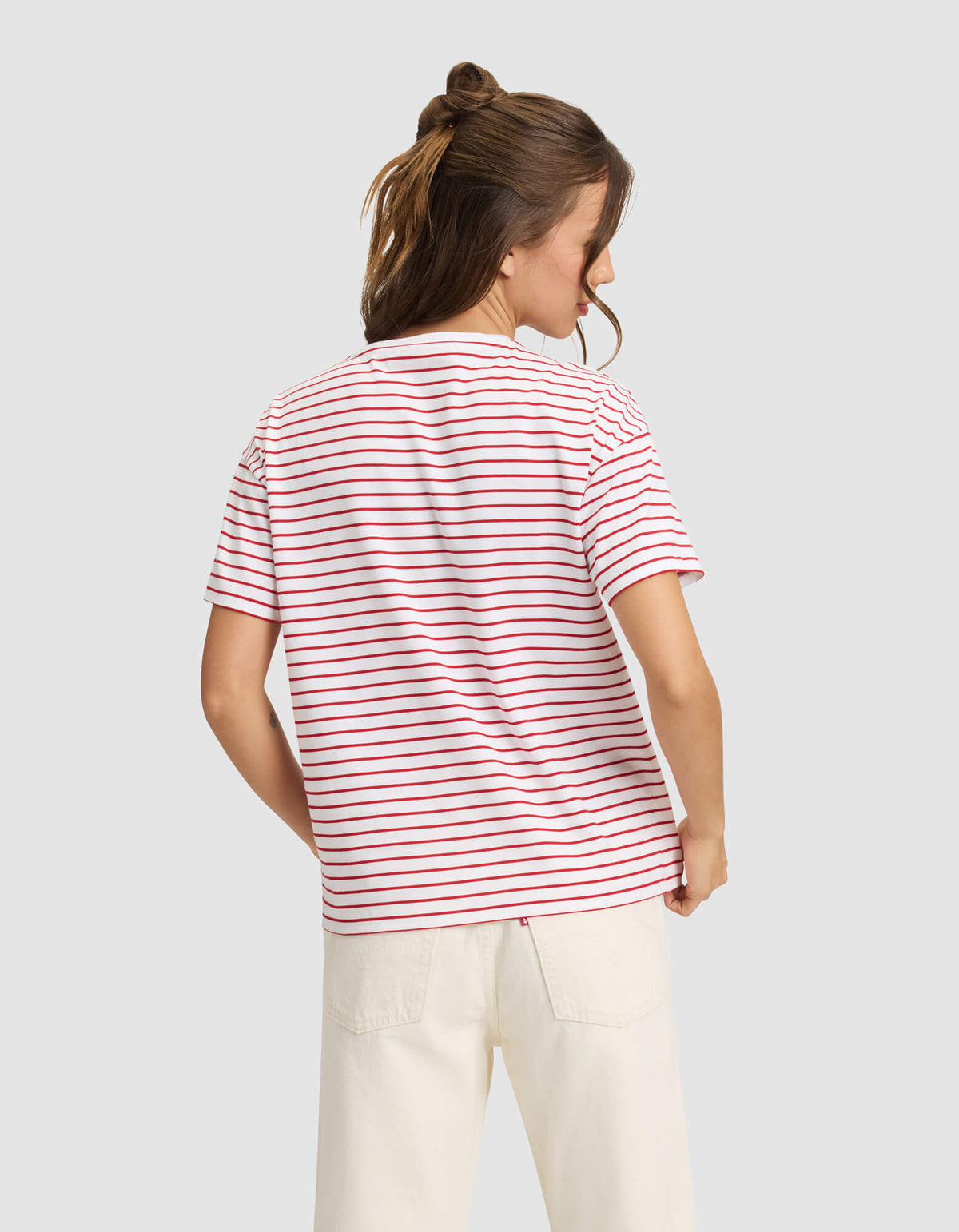 A woman with light brown hair in a half-up style is shown from the back wearing Cozy Earth’s Women’s All Day Tee—white with red and blue stripes—and off-white pants, standing against a plain light gray background. |Color:Crimson Stripe