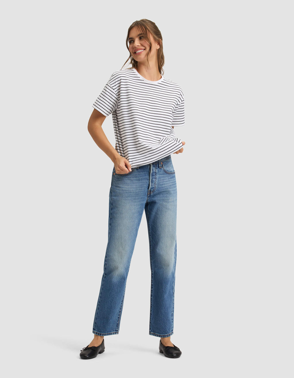 A woman with light brown hair tied back smiles while wearing the Cozy Earth Women's All Day Tee in white and black stripes, paired with blue jeans and black flats. She poses with one hand in her pocket against a plain light gray background. |Color:Jet Black Stripe