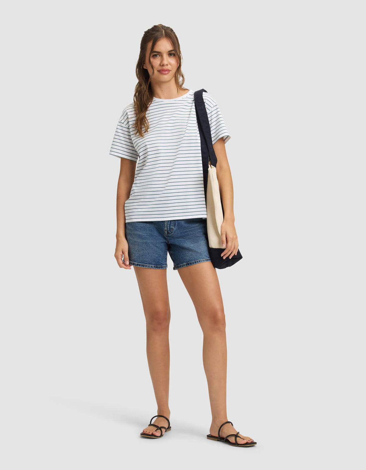 A woman with long, wavy hair stands against a plain background, smiling slightly and wearing the Cozy Earth Women's All Day Tee in white and gray stripes, paired with denim shorts, black sandals, and a navy-beige tote bag over her shoulder. |Color:Oceanside Stripe