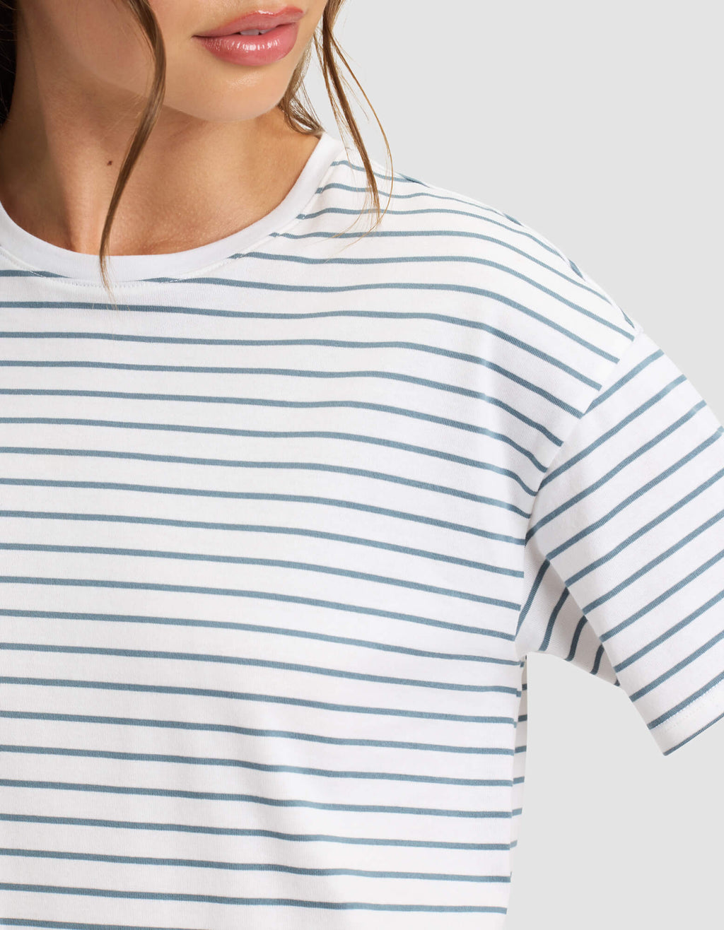 A close-up shows a person wearing Cozy Earth's Women's All Day Tee in white with thin blue horizontal stripes, featuring only the lower face and upper torso against a plain light background. |Color:Oceanside Stripe