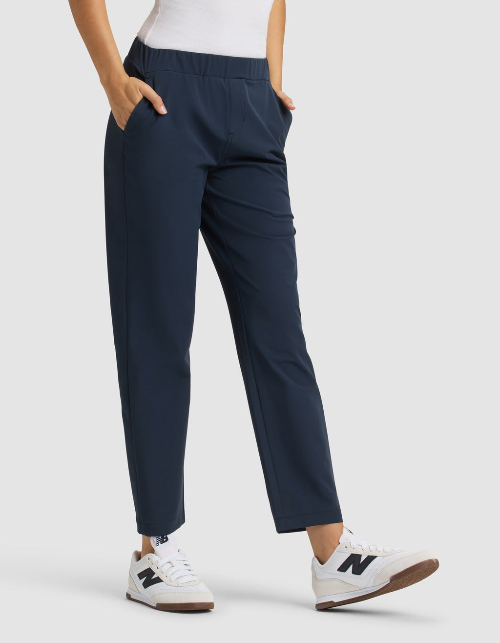 A person wears Cozy Earth Women's Always Cropped Pant in navy blue with a white top and white sneakers with brown soles. Hands are in the pants' pockets, standing against a plain light gray background. |Color:Eclipse