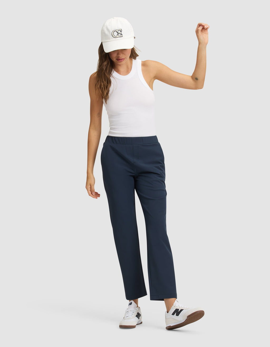 A woman models the Cozy Earth Women's Always Cropped Pant in navy blue, paired with a white sleeveless tank, white sneakers, and a white cap, standing against a light gray background with one arm raised and the other at her side. |Color:Eclipse