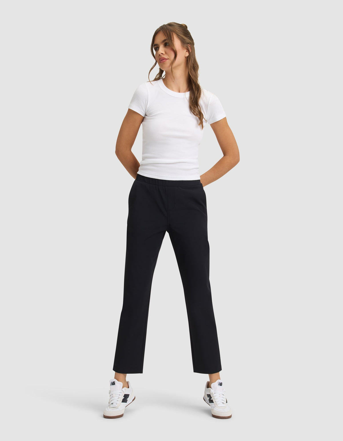A woman stands against a light gray background, smiling with her hands in her pockets, wearing Cozy Earth's Women's Always Cropped Pant in black, a fitted white t-shirt, and white sneakers with blue and tan accents. |Color:Jet Black