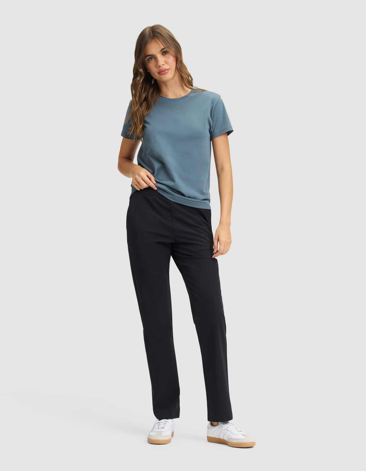 A woman with long, wavy hair poses against a plain background, wearing Cozy Earth's Women's Always Pant in black, paired with a blue short-sleeve t-shirt and white sneakers. One hand is in her pocket. |Color:Jet Black